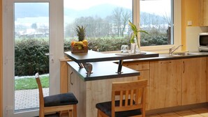 A kitchen with wooden cabinets, a black countertop, and a view of a snowy landscape through large windows. - Ferienwohnung am Rain (Winden im Elztal)