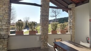 Een overdekt terras met stenen zuilen, een houten tafel en witte stoelen.
