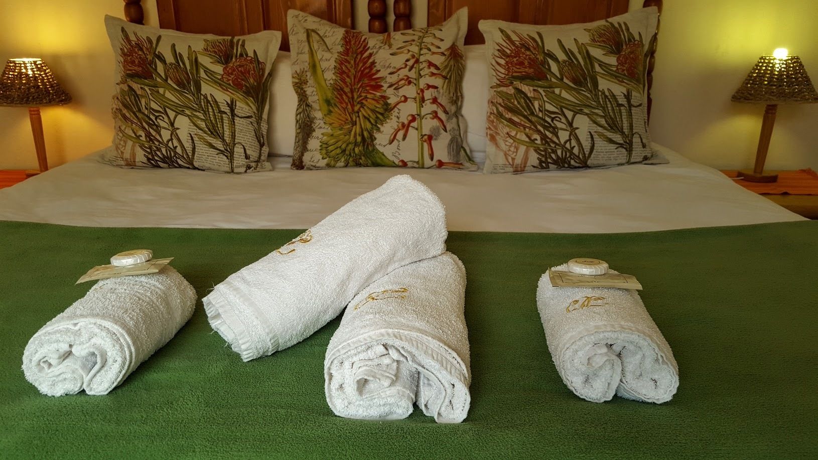 A neatly made bed with floral pillows, a green bedspread, and three rolled towels with gold tags.