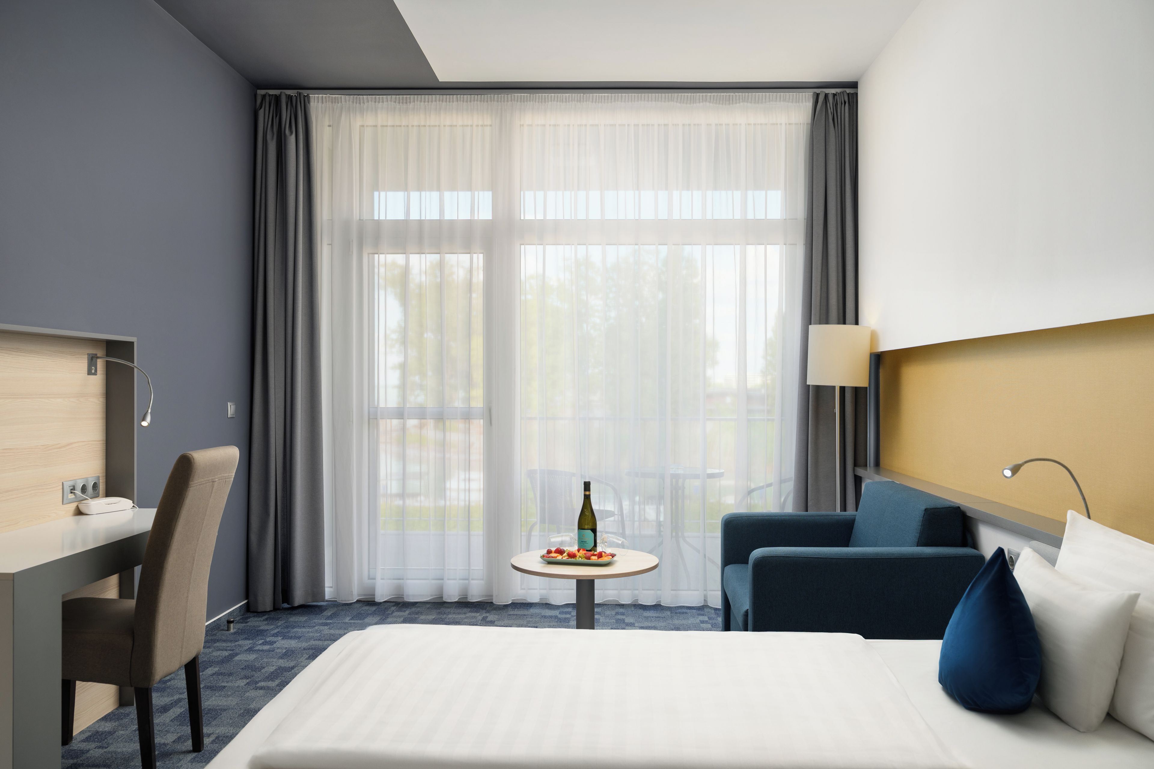 A hotel room with a bed, a desk, a chair, a small table with a bottle and fruit, and a window with curtains.