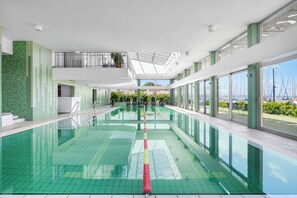 A modern indoor swimming pool with clear water, a starting block, and a view of the sea through large windows.
