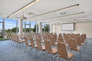 Un salón de conferencias con filas de sillas, una pantalla de proyector y grandes ventanales que ofrecen vistas a árboles y edificios.