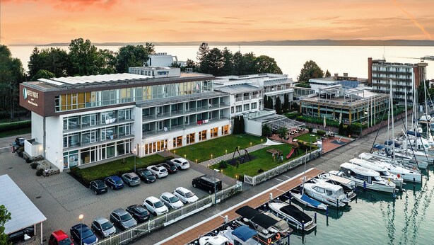 A modern hotel with a glass facade, a parking lot, and a marina with boats.
