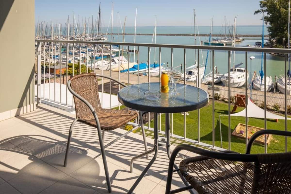 Un balcón con sillones de mimbre, una mesa de vidrio y vista a un puerto deportivo con barcos.
