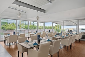 Un restaurant avec des tables dressées pour le service, de grandes fenêtres donnant sur un paysage verdoyant et un plan d’eau.