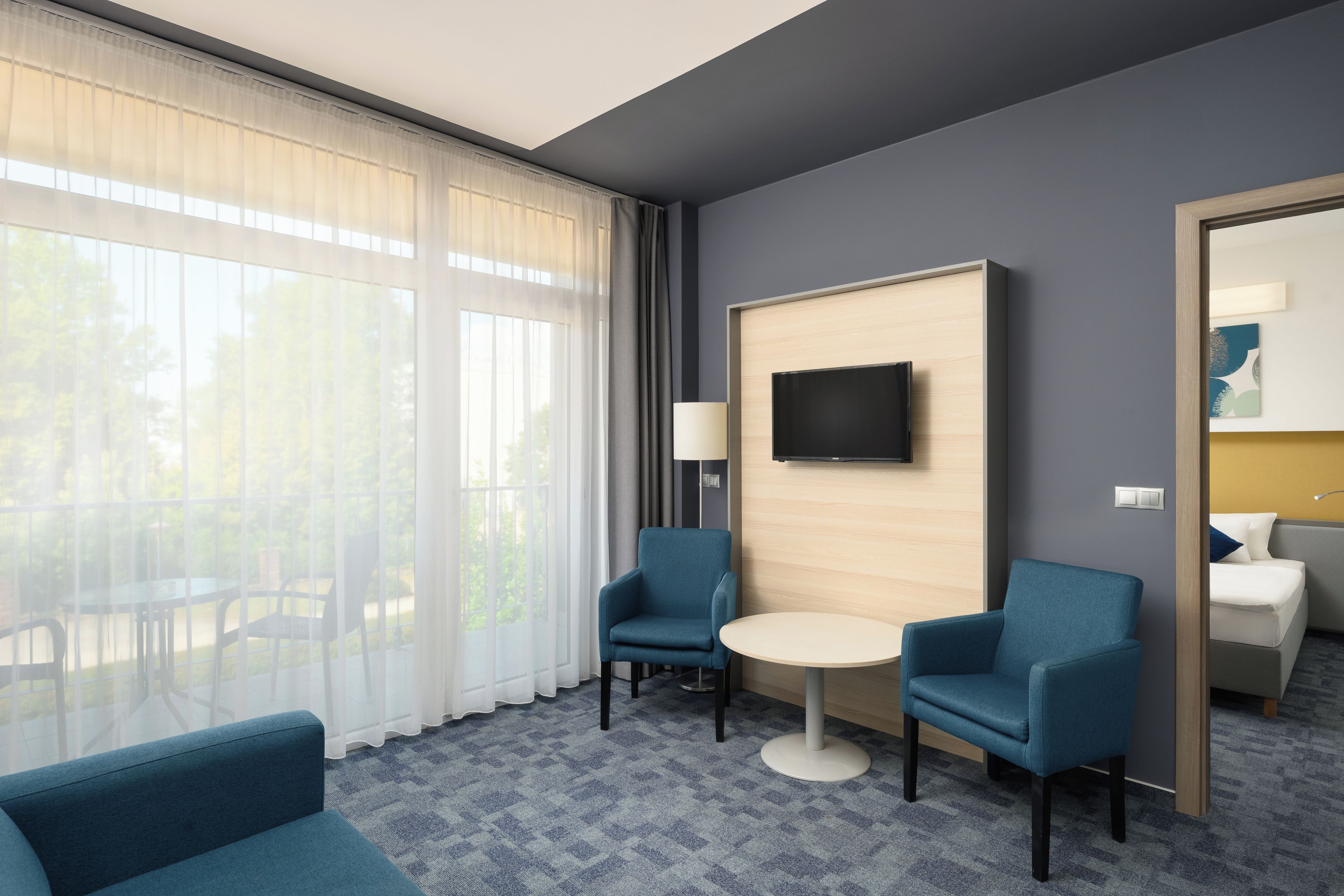A hotel room with a large window, two armchairs, a small round table, a flat-screen TV mounted on a wooden panel, and a bed with a nightstand.