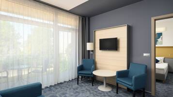 A hotel room with a large window, two armchairs, a small round table, a flat-screen TV mounted on a wooden panel, and a bed with a nightstand.