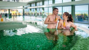 A couple in a large indoor swimming pool with clear views of the cityscape through large windows.