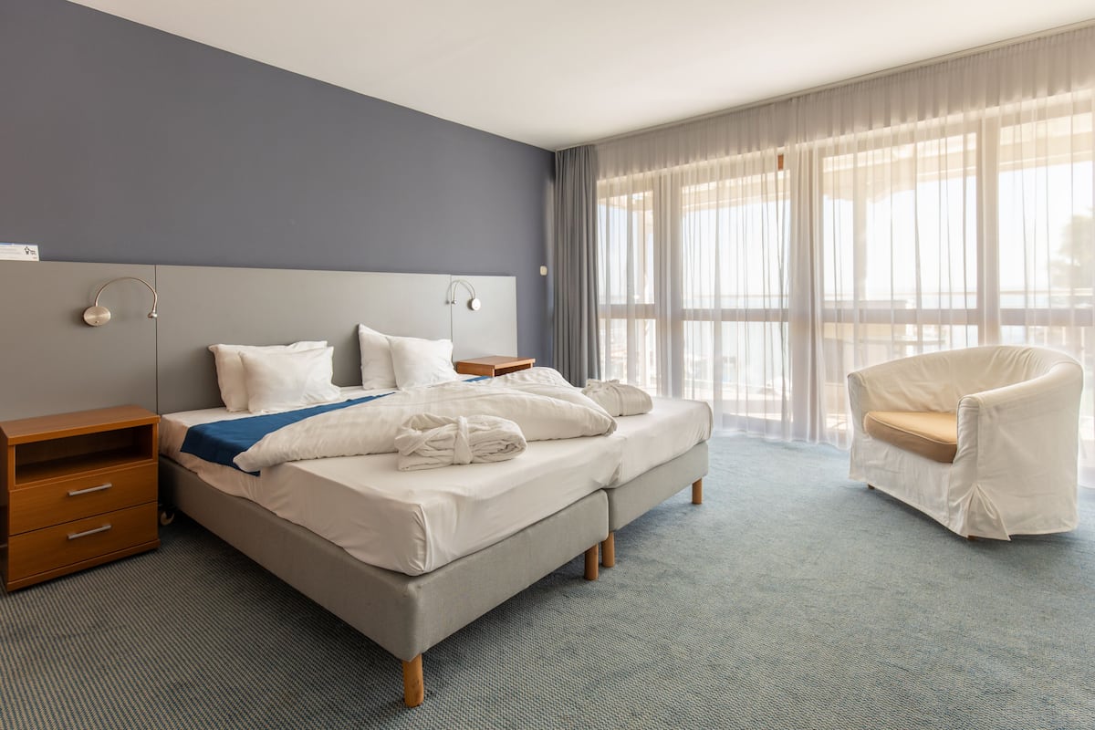Una habitación de hotel con una cama grande, una mesita de noche, una silla y una ventana con cortinas traslúcidas.