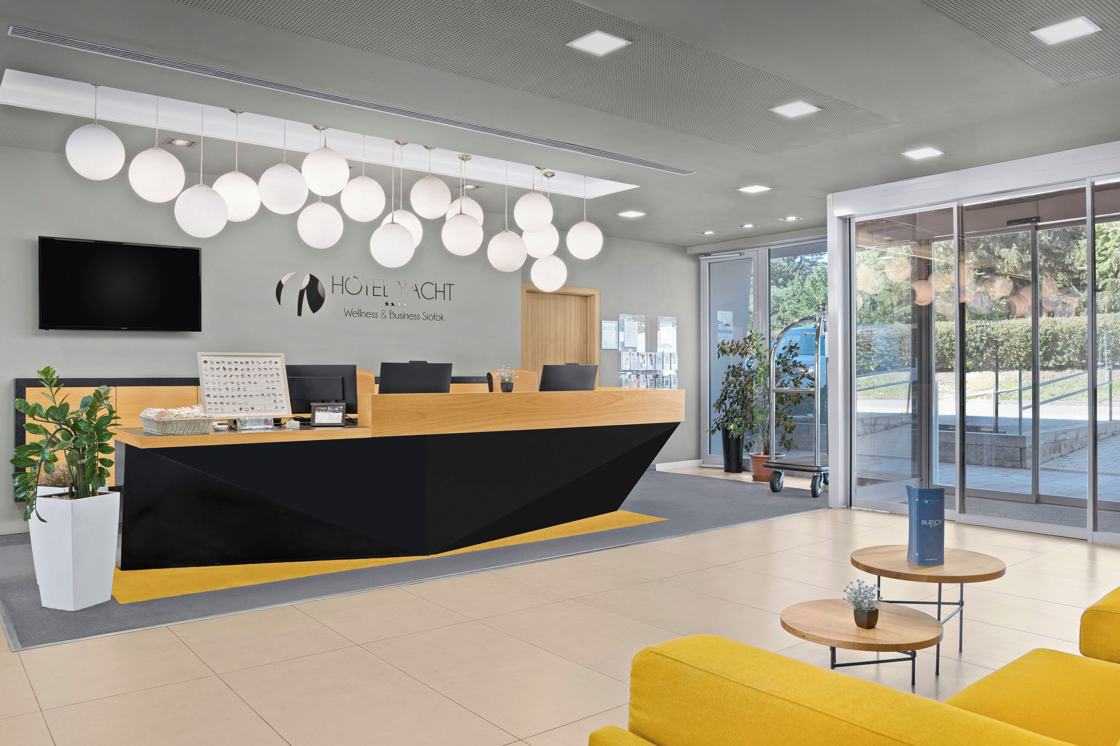 Un mostrador de recepción de hotel con diseño moderno, un televisor de pantalla plana y el logotipo de 'HOTEL YACHT Wellness & Business Söfok' en la pared.