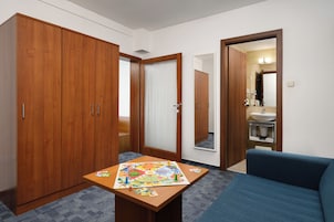 Quarto de hotel com guarda-roupa de madeira, uma pequena mesa com um jogo de tabuleiro, um sofá azul e um banheiro visível através de uma porta aberta.