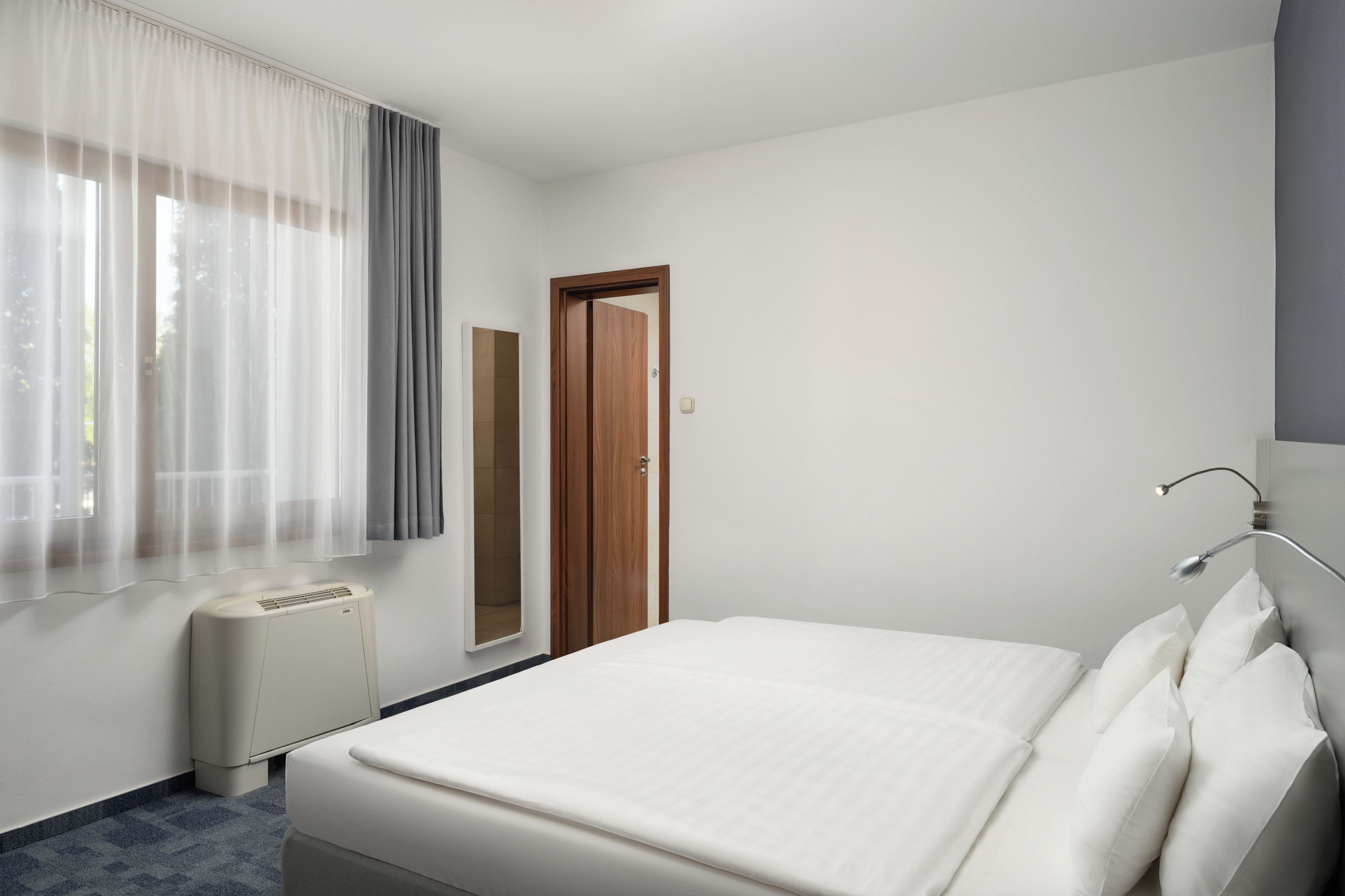 Habitación de hotel con cama, ventana con cortinas, puerta, espejo y lámpara de noche.