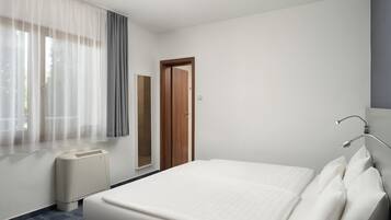 Habitación de hotel con cama, ventana con cortinas, puerta, espejo y lámpara de noche.