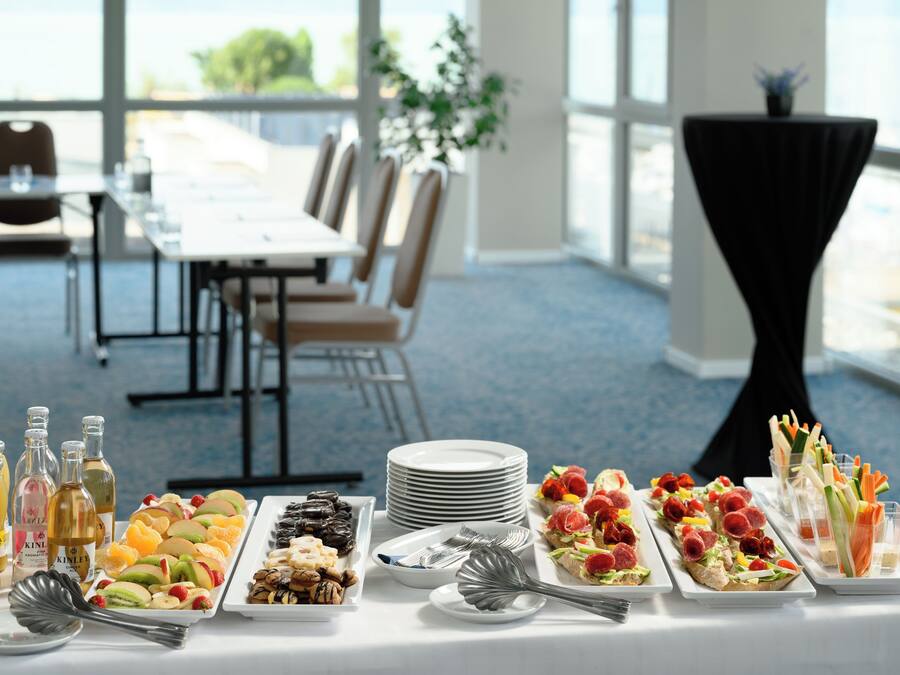Une table de buffet proposant divers plats et accompagnements, un coin repas avec des chaises et une table, et une vue sur l’extérieur grâce à de grandes fenêtres.
