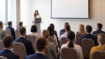 Un salón de conferencias con un orador en un atril, un público sentado frente a él y una pantalla de proyección.