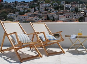 Two wooden lounge chairs with blue and white striped cushions on a rooftop terrace with a city view in the background. - Hydras Chromata (Hydra)