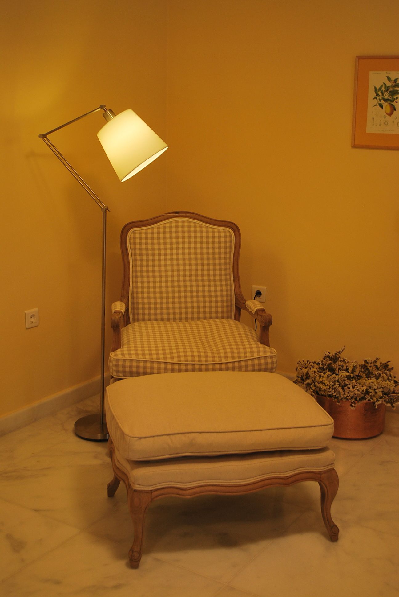 Un fauteuil à motifs à carreaux, accompagné d’un pouf assorti, une lampe sur pied et une plante en pot, dans une pièce aux murs jaunes.