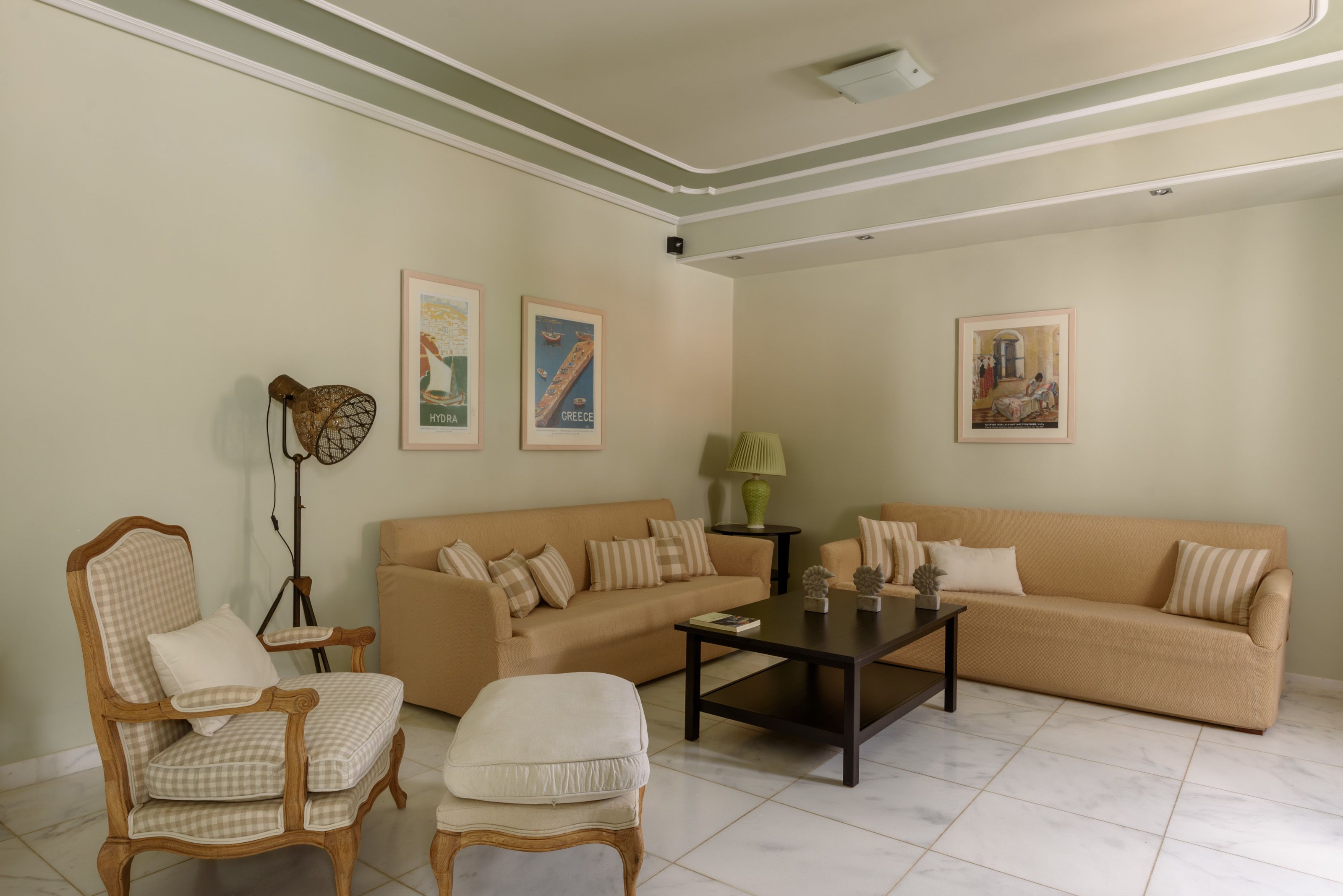 A living room with a beige sofa, two armchairs, a coffee table, and a floor lamp. There are three framed pictures on the wall.