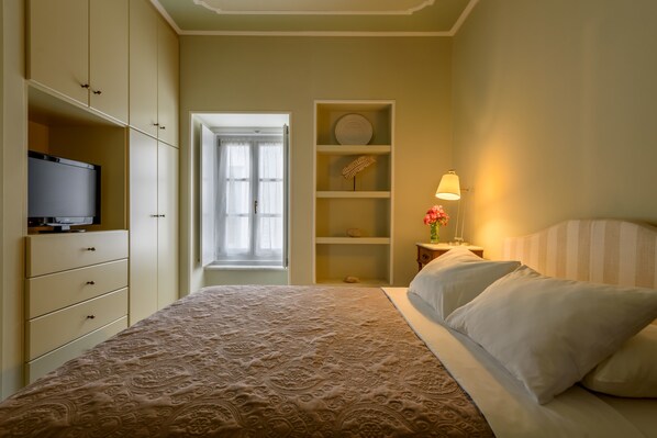 Un dormitorio con una cama, un televisor sobre un mueble, una estantería con objetos decorativos y una lámpara.