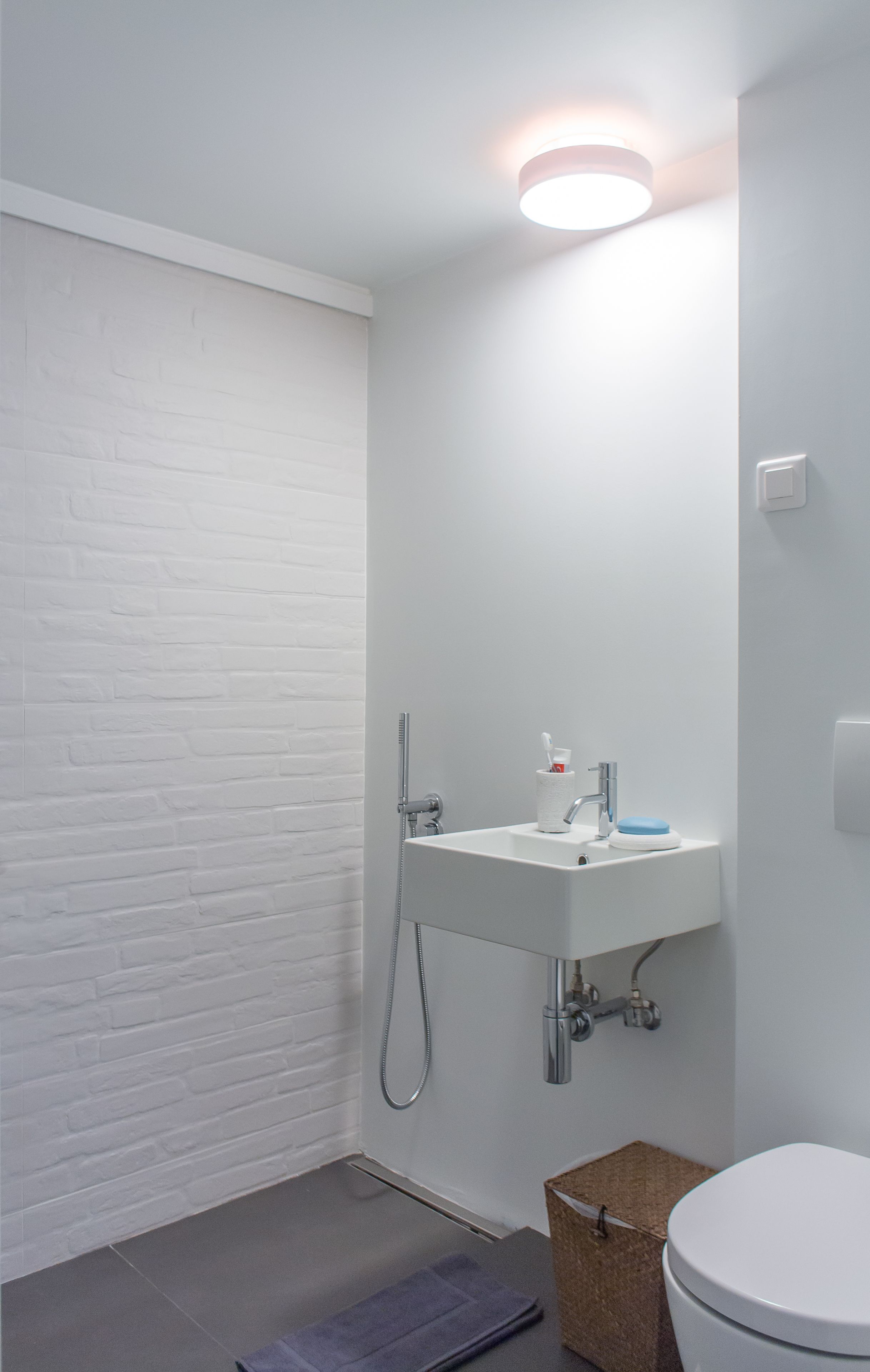A small, modern bathroom with a white brick wall, a white sink with a blue soap dish, a shower, and a white toilet.