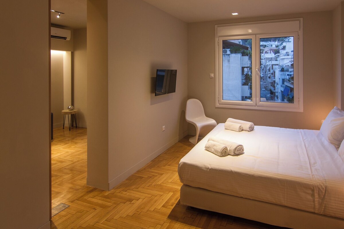 A bedroom with a bed, a chair, a TV mounted on the wall, and a window with a view of buildings.