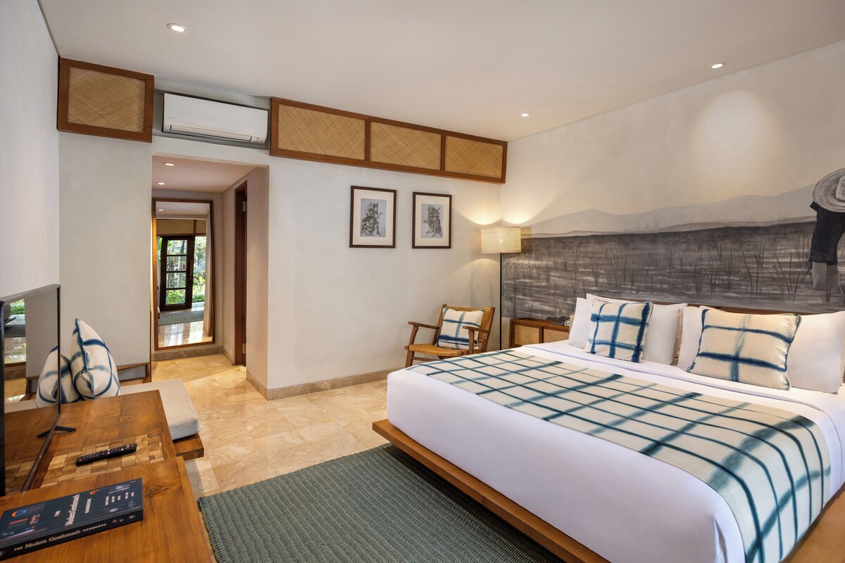 A modern bedroom with a large bed, a desk with a computer, and a chair. There are framed pictures on the wall and a painting above the bed.