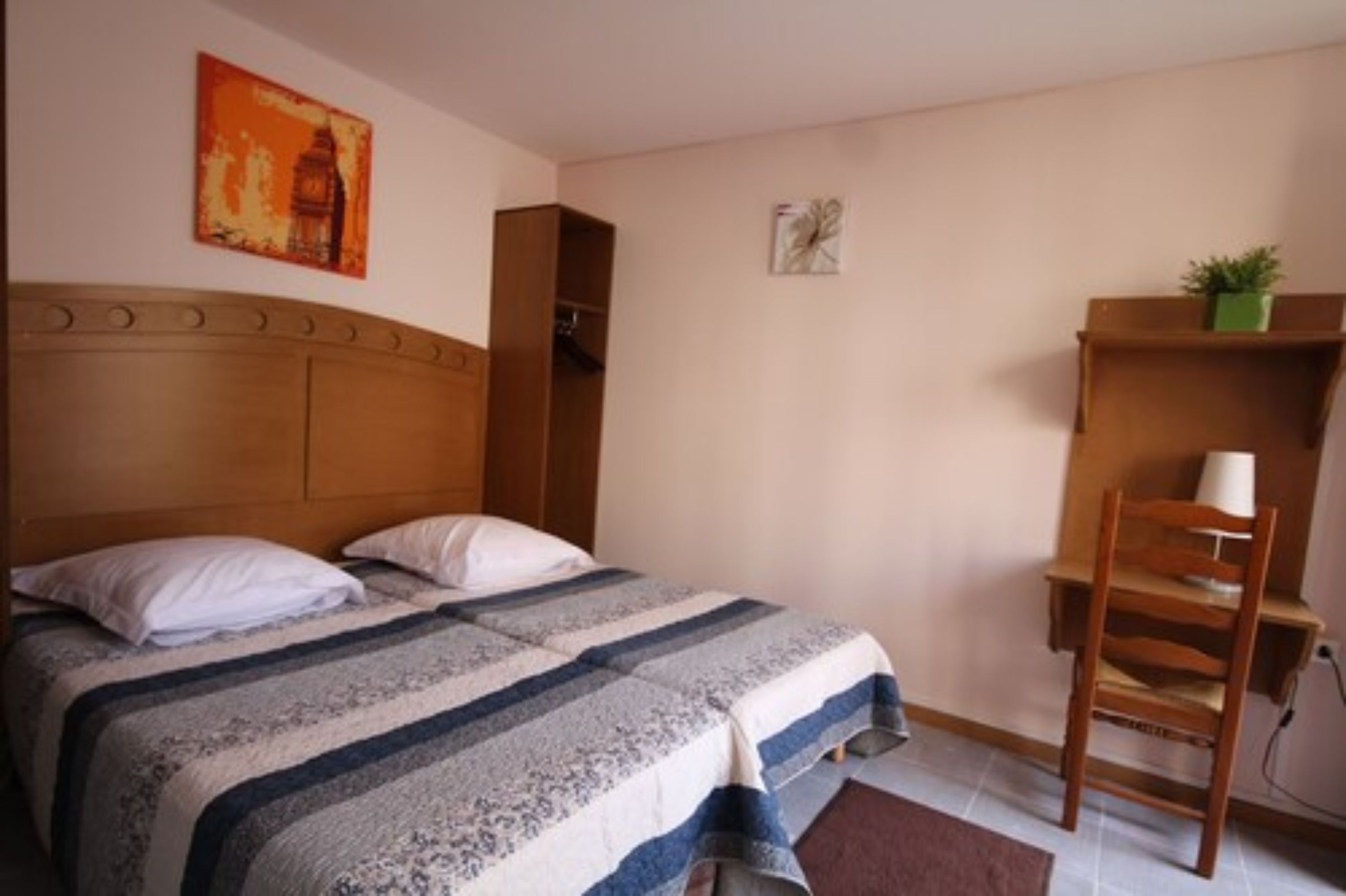Un dormitorio con cabecera de madera, cama con manta a rayas, una silla, estante con una planta y un cuadro en la pared.