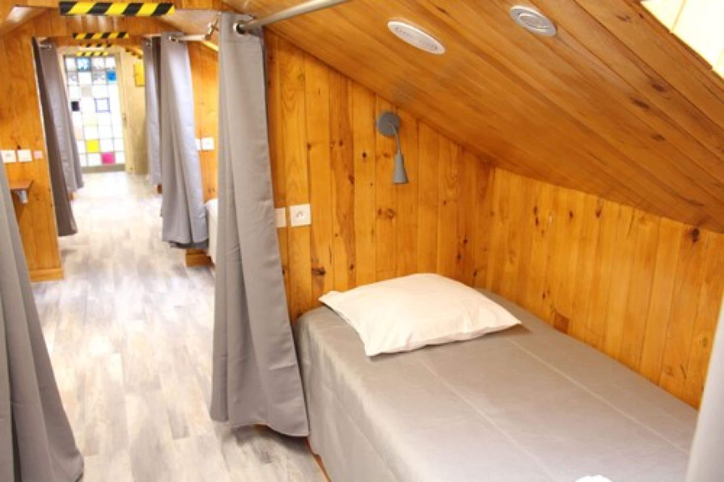 A small, single-occupancy room with wooden walls, a bed with a white pillow, and a ceiling fan.