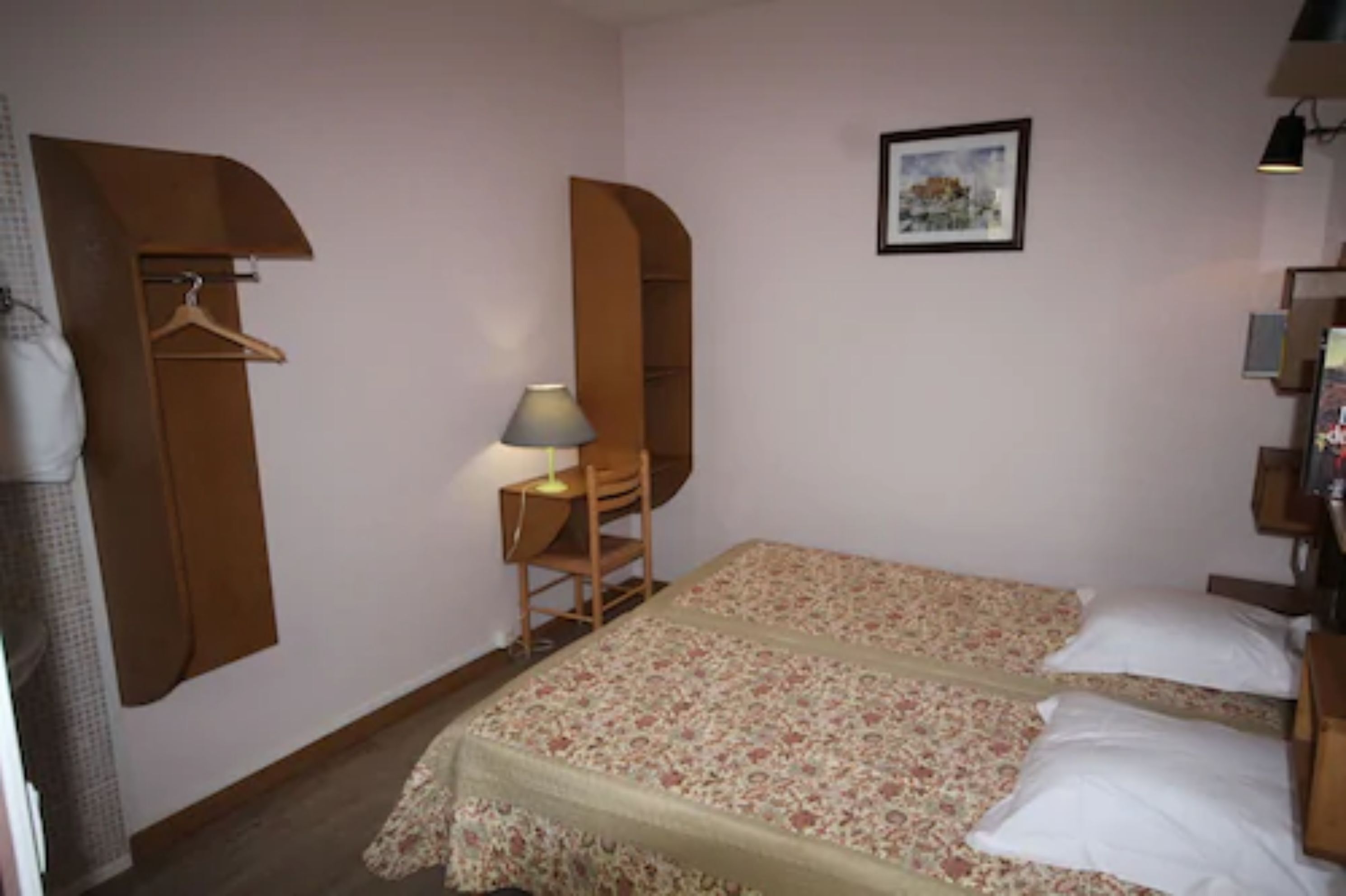 Une petite chambre avec deux lits simples, une table de chevet, une chaise, une lampe, une armoire et un tableau encadré au mur.