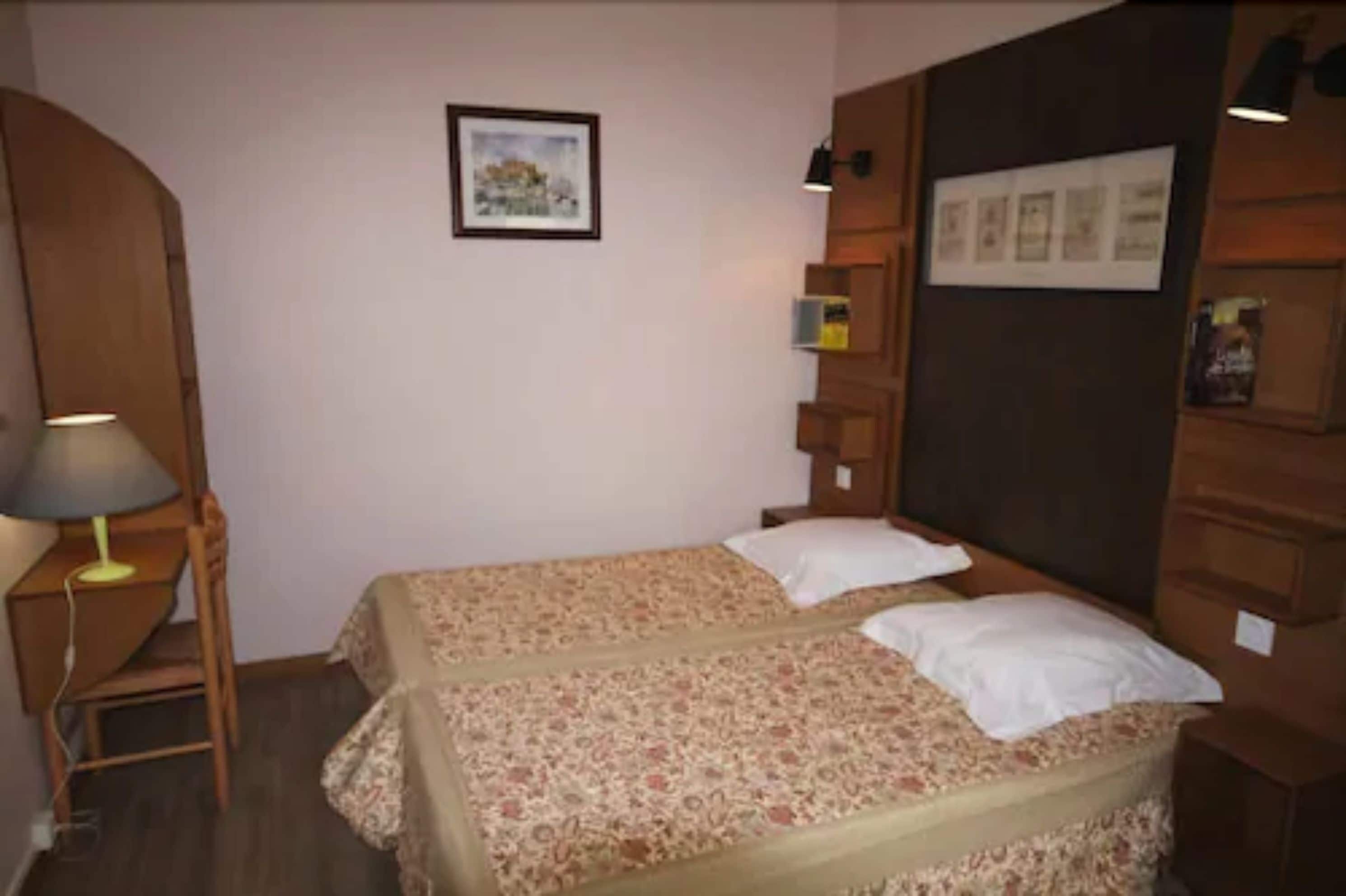 A bedroom with two beds, a nightstand, a lamp, and a wall-mounted shelf.