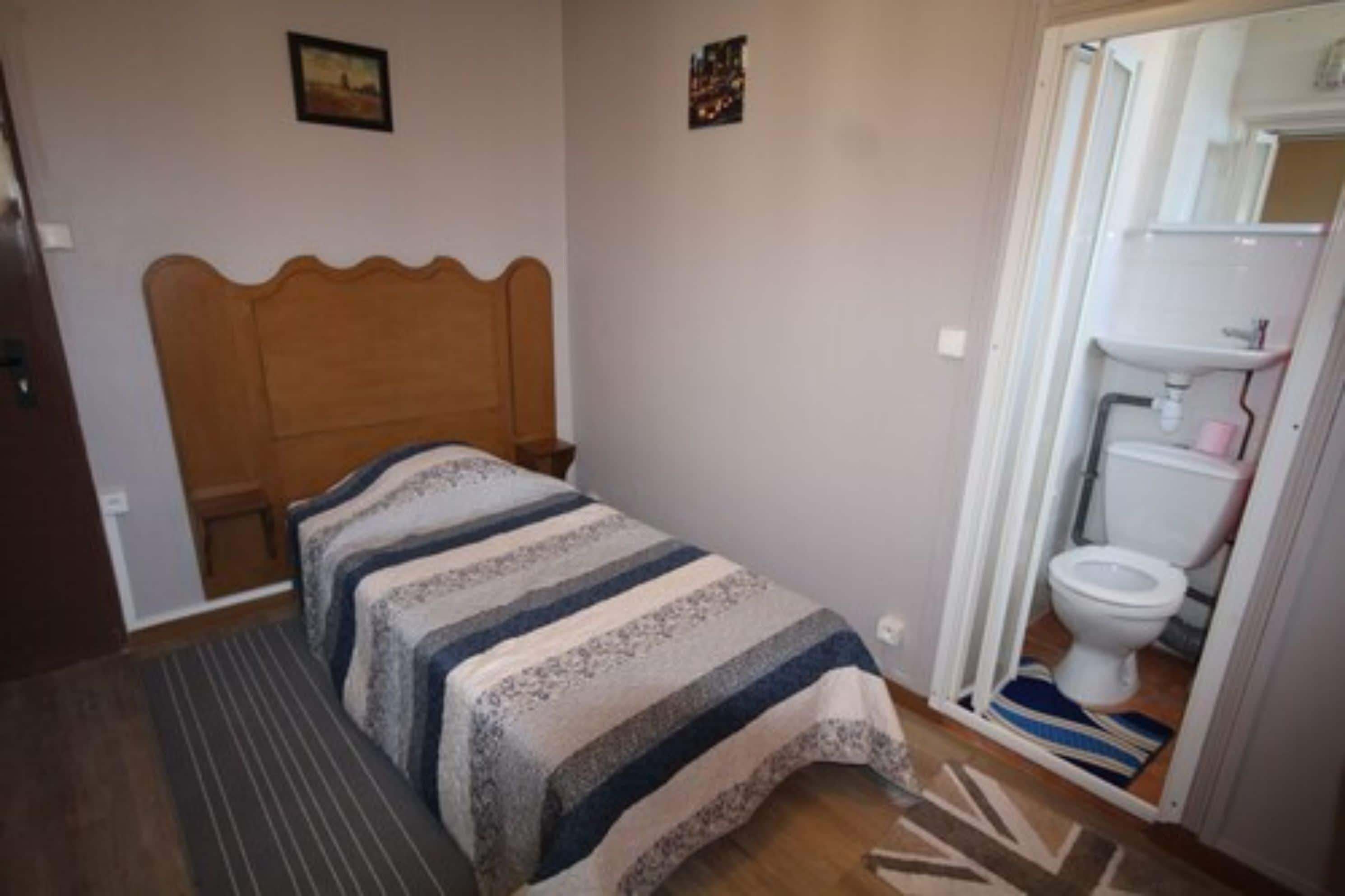 Habitació petita amb un llit individual, capçalera de fusta, una nitzeta, taula de nit, una finestra petita i un bany amb vàter i rentamig.