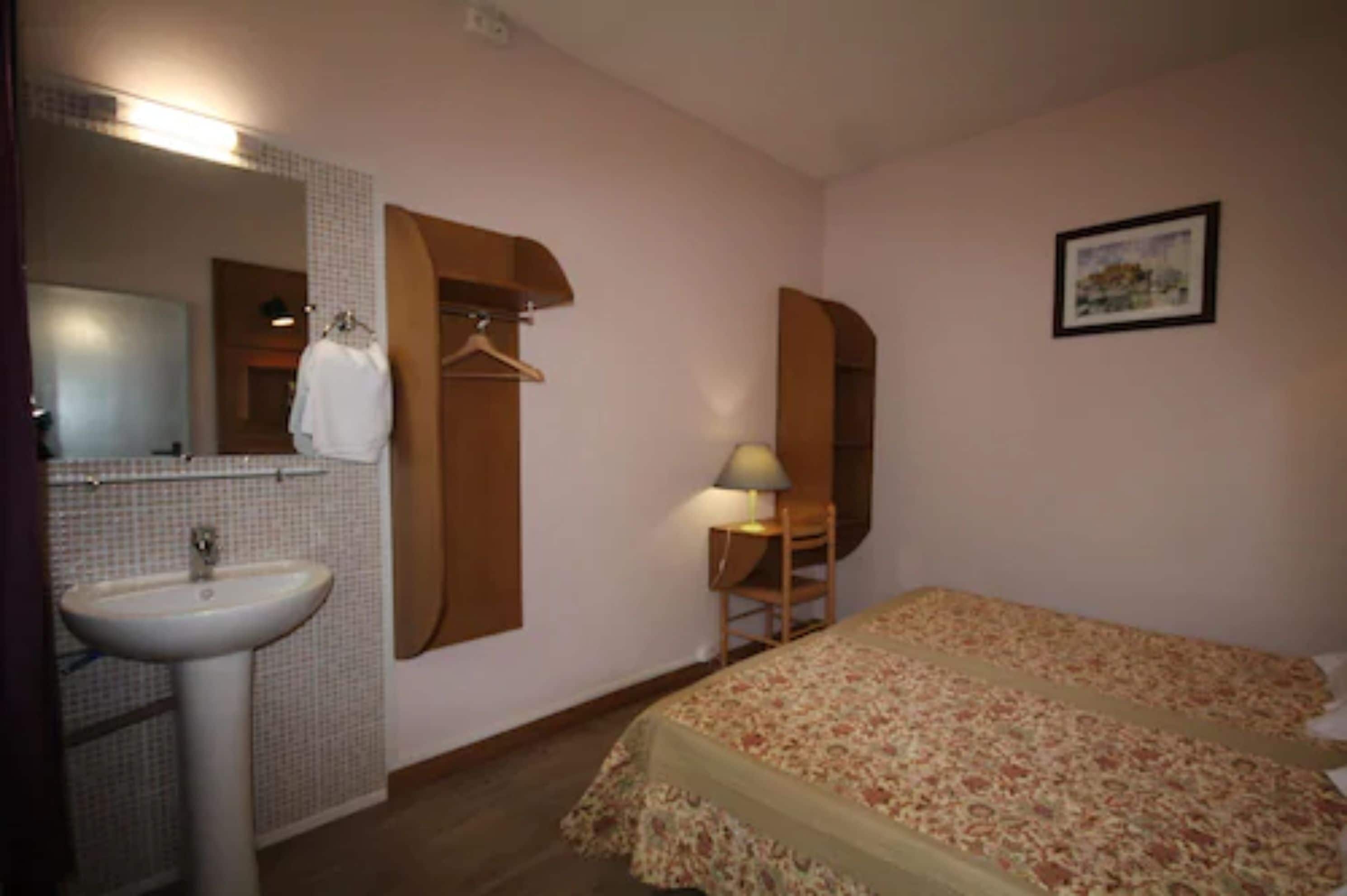 Een slaapkamer met een bed, een nachtkastje met lamp, een kapstok en een badkamer met wastafel en spiegel.