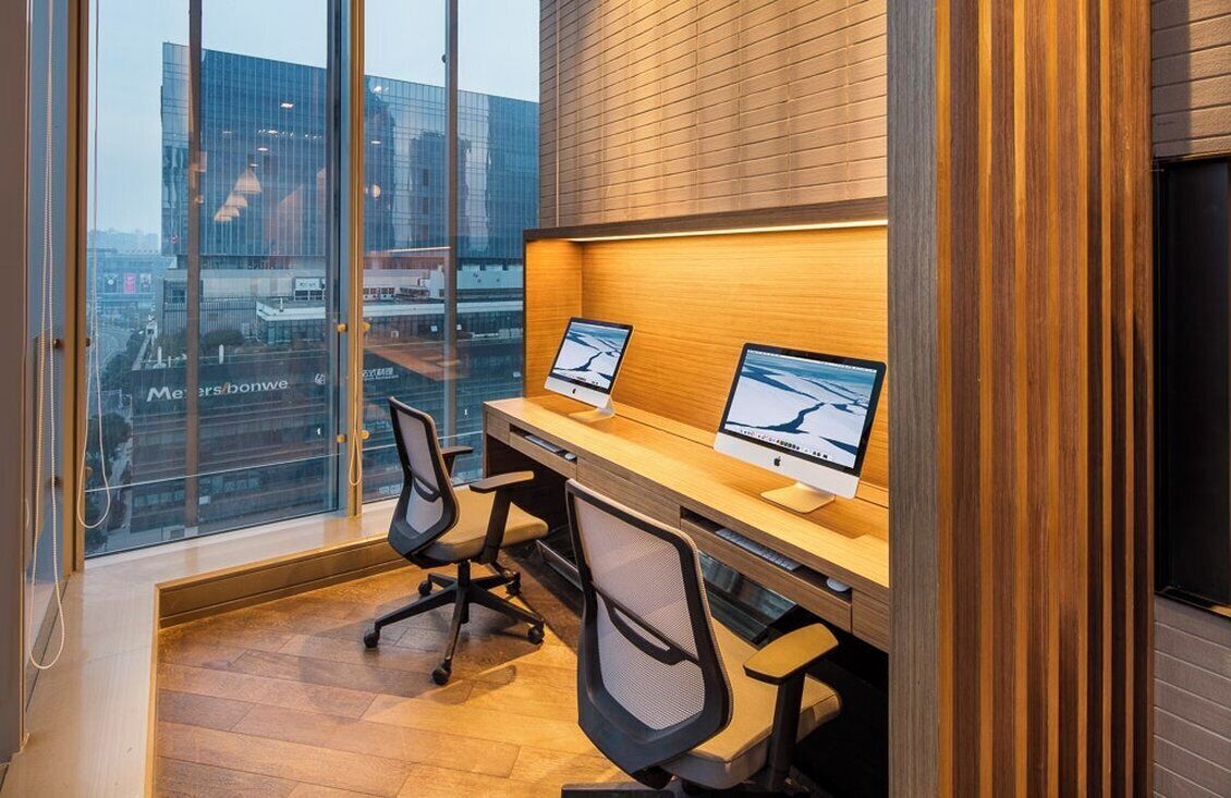 A modern office space with two desks, each with a computer monitor, office chairs, and a large window offering a city view.