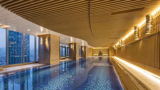 A modern indoor swimming pool with a tiled floor, wooden paneling on the walls, and a view of city buildings. - Ascott Central Wuxi (Wuxi)