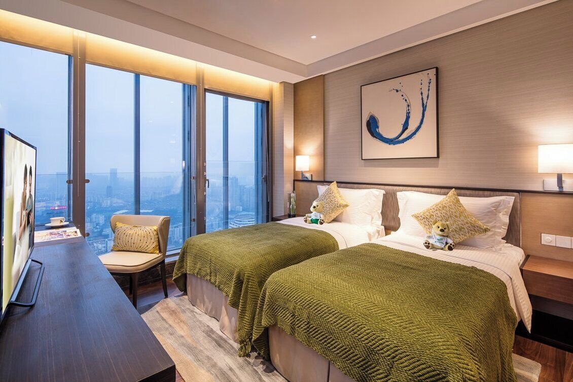 A hotel room with two beds, a desk, a chair, and a large window offering a city view.