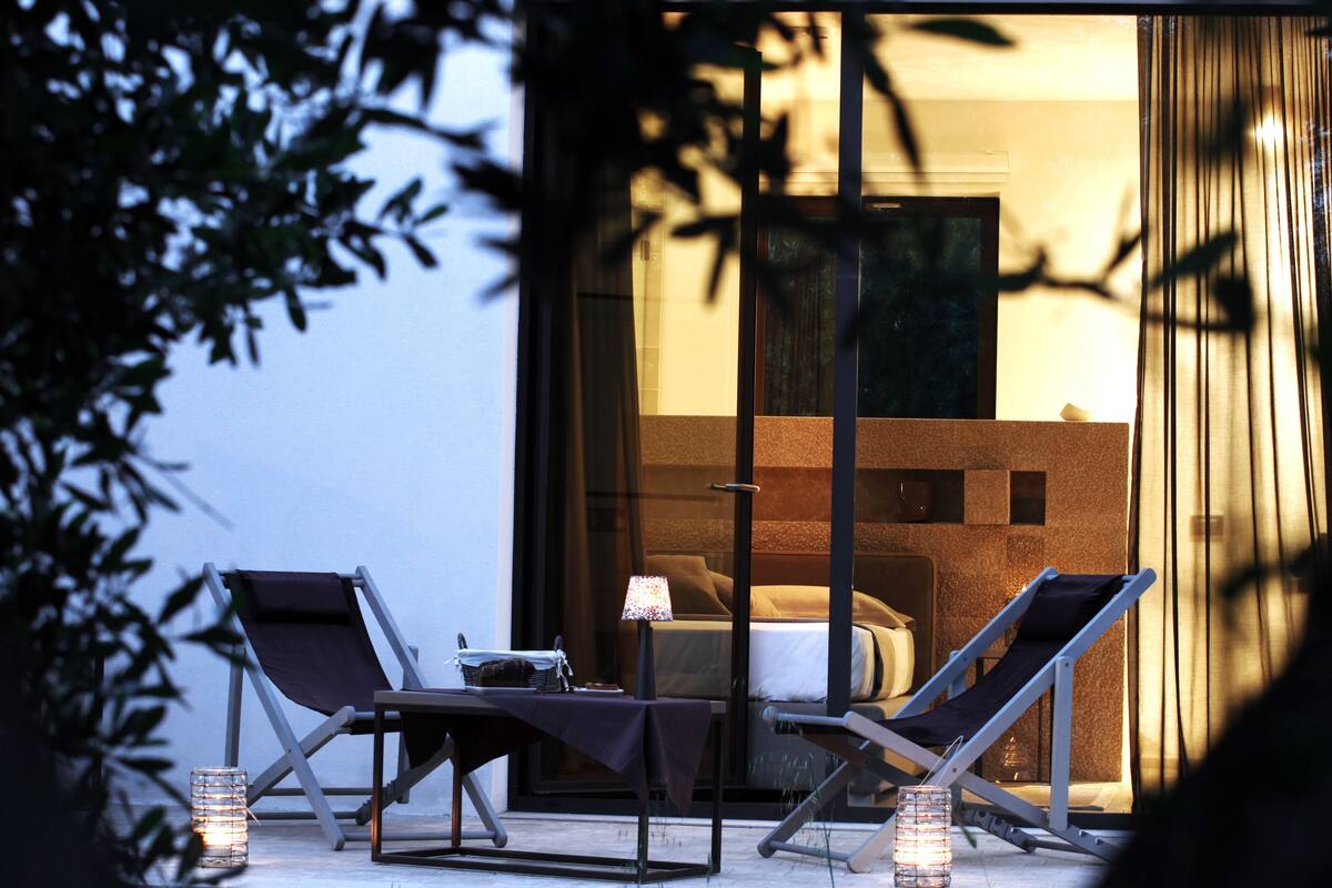 Un área de estar al aire libre con dos sillones y una mesa, una vela de cristal, y vista a un dormitorio a través de una puerta de cristal.
