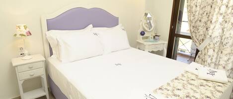 Una cama bien hecha con sábanas blancas y toallas de estampado floral, una mesita de noche con lámpara, y una ventana con cortinas estampadas con flores.