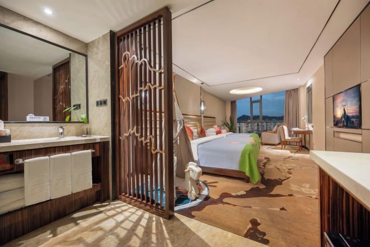 A hotel room with a bathroom, a bed with a green blanket, a cowhide rug, a wooden partition, and a city view.