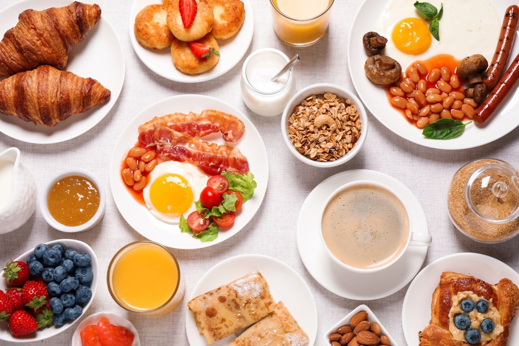 A breakfast spread with croissants, eggs, bacon, beans, sausages, and various fruits and beverages.