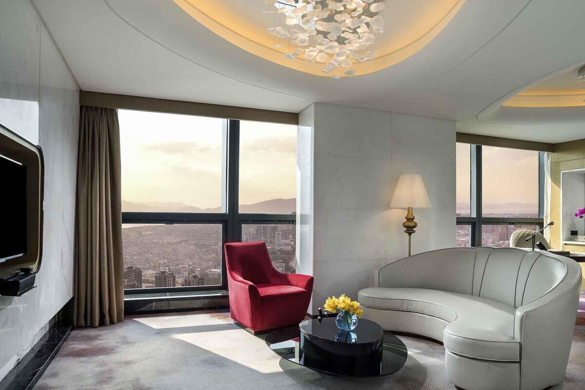 A modern hotel room with a curved white sofa, a red armchair, a black coffee table with flowers, a flat-screen TV, and large windows offering a city view.