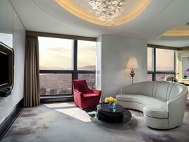 Una camera d'albergo moderna con un divano bianco curvo, una poltrona rossa, un tavolino nero con fiori, una televisione a schermo piatto e ampie finestre con vista sulla città.