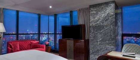 A hotel room with a large bed, a desk, a television, and a view of a cityscape at night.