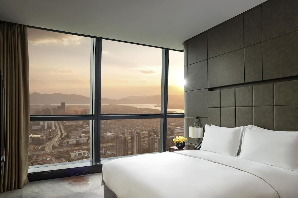 A hotel room with a large bed, a nightstand with a lamp, and a view of a cityscape through large windows.