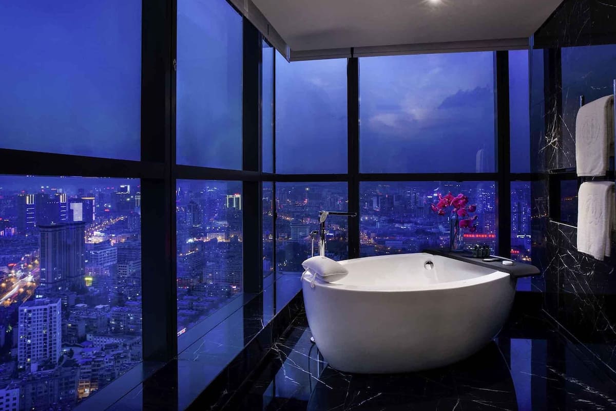 A modern bathroom with a large bathtub, a view of a cityscape through floor-to-ceiling windows, and marble flooring.