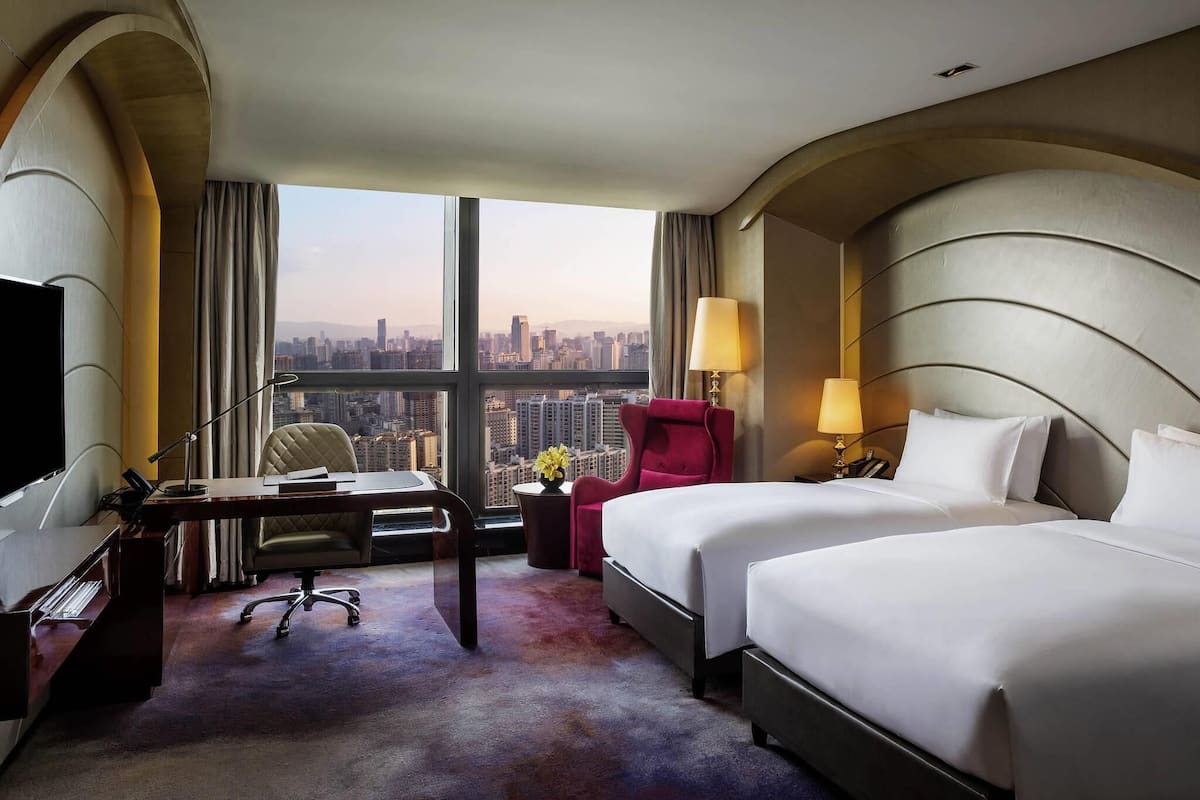 A hotel room with a desk, two beds, a chair, a lamp, and a view of the cityscape.