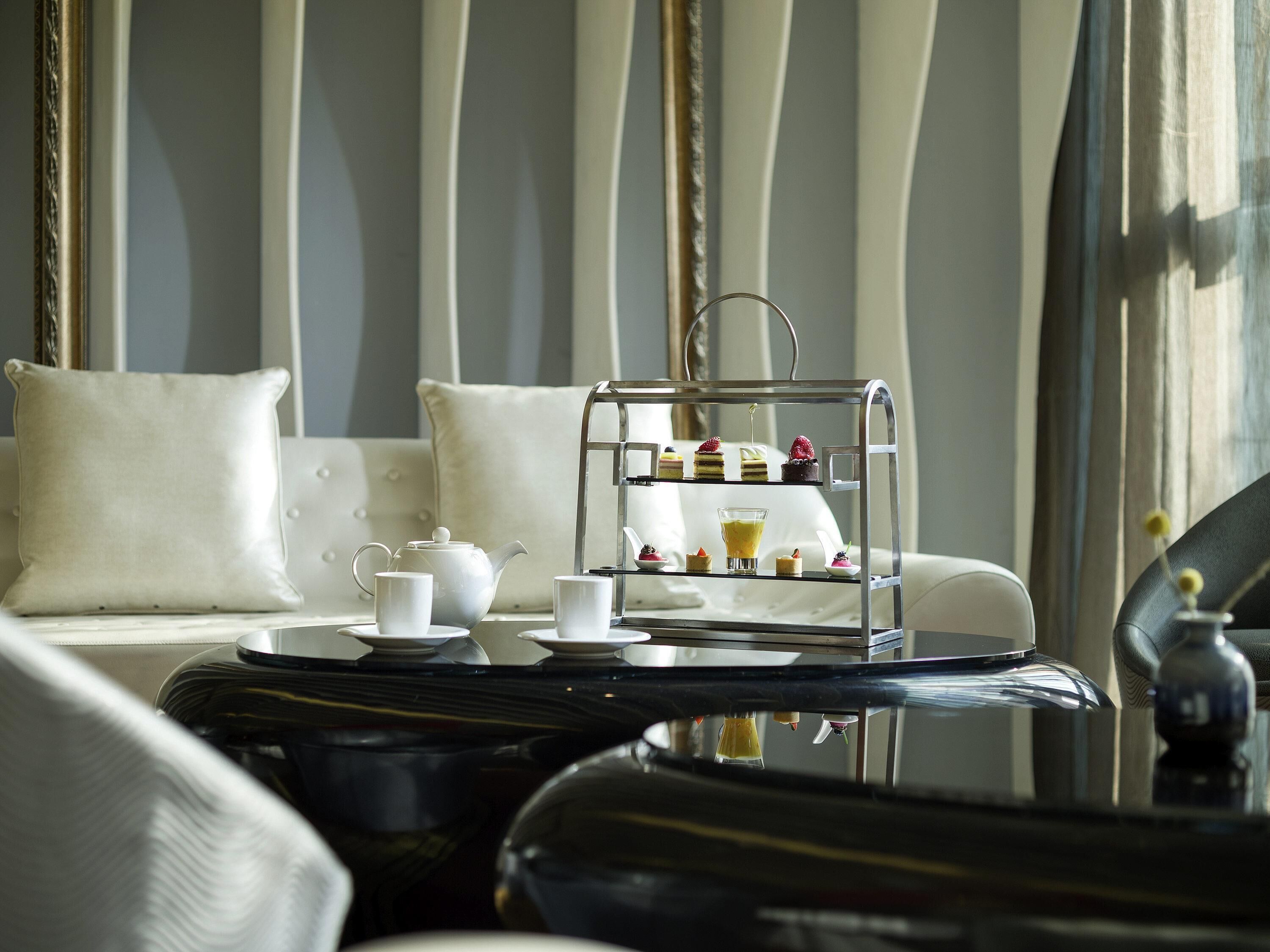 A three-tiered dessert cart with various pastries and a teapot on a table in a room with a sofa and curtains.