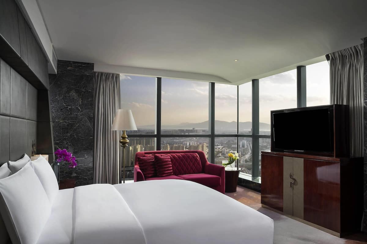 A hotel room with a large bed, a red sofa, a TV, and a city view through large windows.