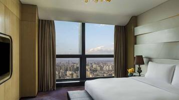 A hotel room with a large bed, a bench, a TV, a lamp, and a view of a cityscape through a large window.