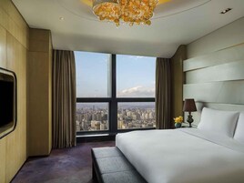 Habitación de hotel con una cama grande, un banco, un televisor, una lámpara y vistas a un paisaje urbano a través de un ventanal.
