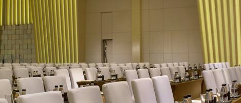 A conference room with rows of white chairs, a long table with water bottles and glasses, and a chandelier.